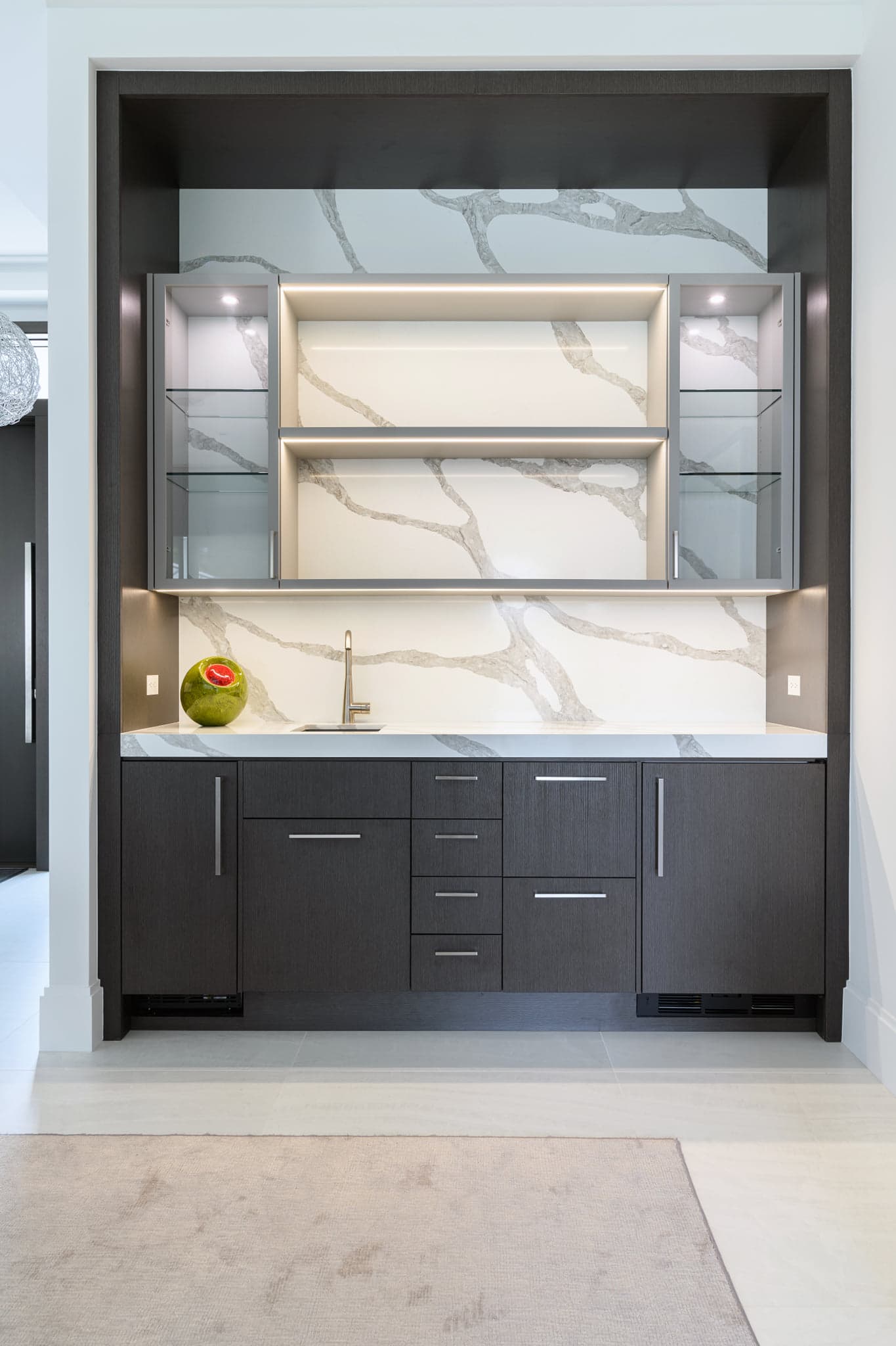 Wet bar with dark oak cabinetry, a Calacatta quartz waterfall backsplash, and lit glass display cabinets.
